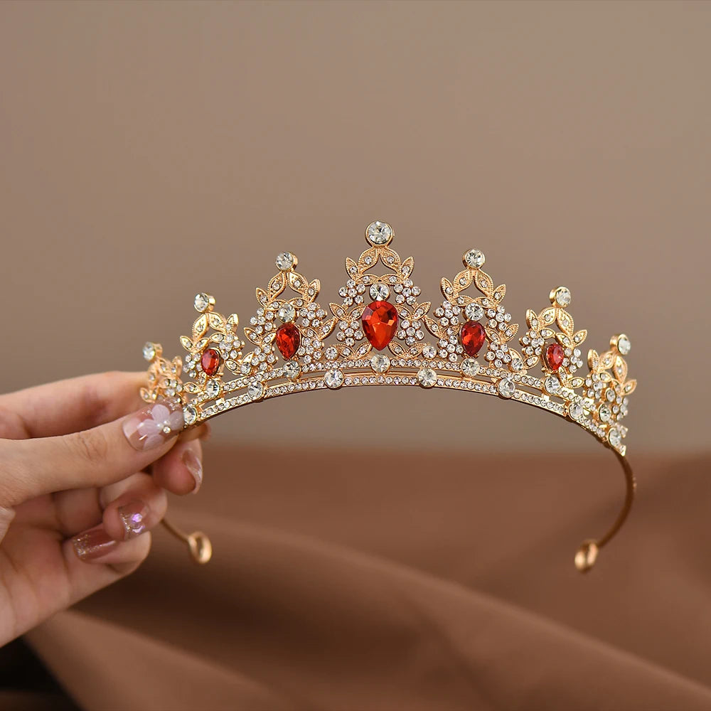 A person holds a golden tiara adorned with sparkling clear crystals and large red gemstones, set against a soft brown background, showcasing intricate detailing.