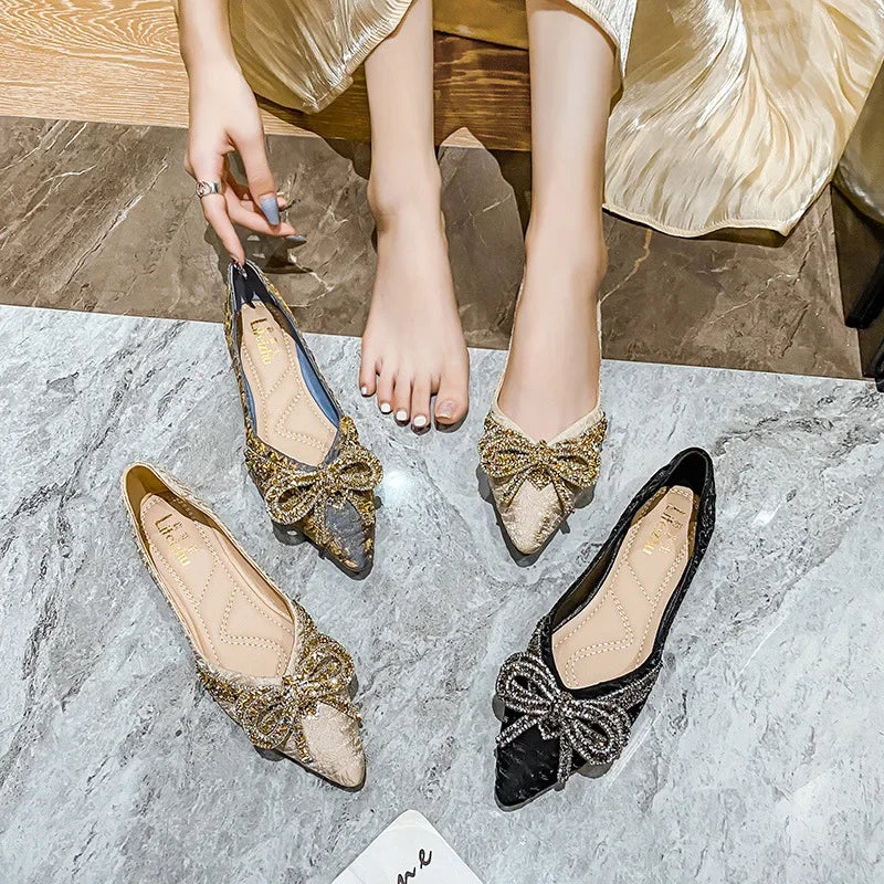 Four embellished ballet flats with large bows on a marble floor. A person wearing cream pants reaches to pick one up, showing a painted toenail.