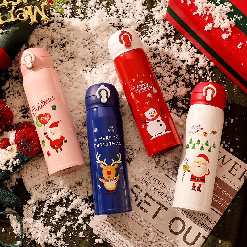 Four festive thermos bottles on snow, each with Christmas designs: pink with Santa, blue with reindeer, red with snowman, and white with Santa and trees, next to a gift box.