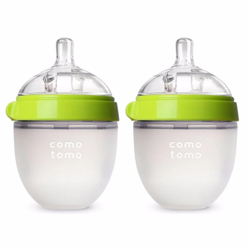 Two Comotomo baby bottles with green lids and clear silicone nipples. The bottles are made of soft, squeezable material with the brand name visible on the front.