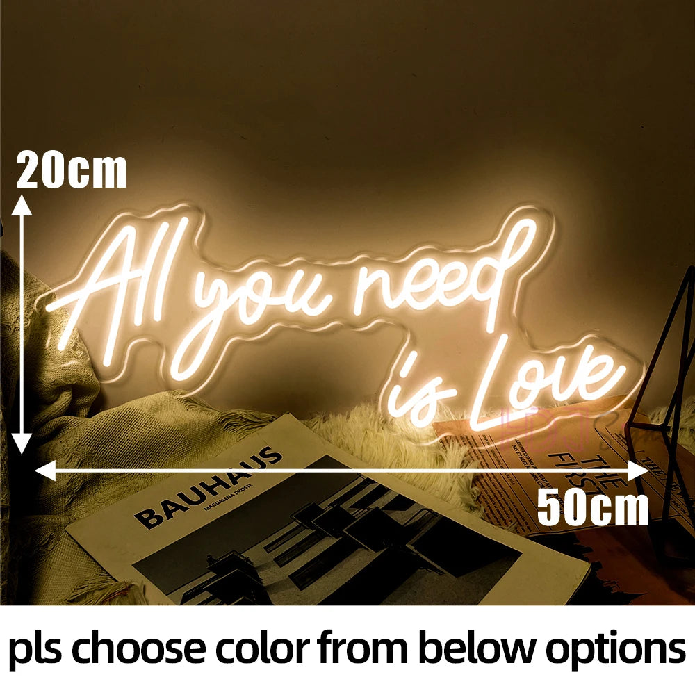 Neon sign reading "All you need is Love" in warm light, measuring 20cm by 50cm. It rests on a soft surface alongside a magazine titled "Bauhaus."