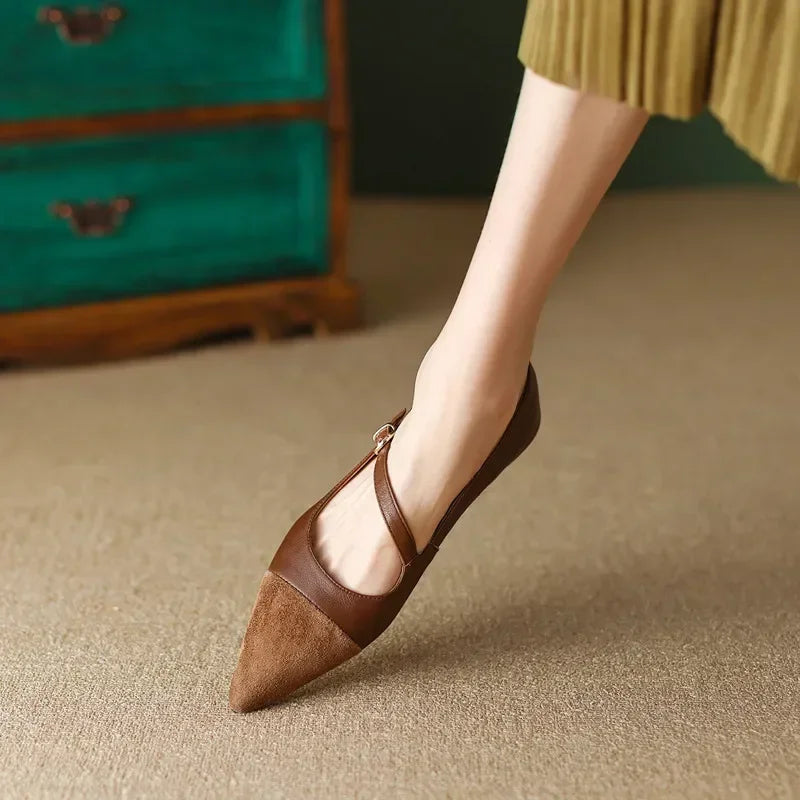 A person wearing a brown pointed toe flat shoe with a strap, standing on a beige carpet. The background features a teal dresser and a brown pleated skirt.