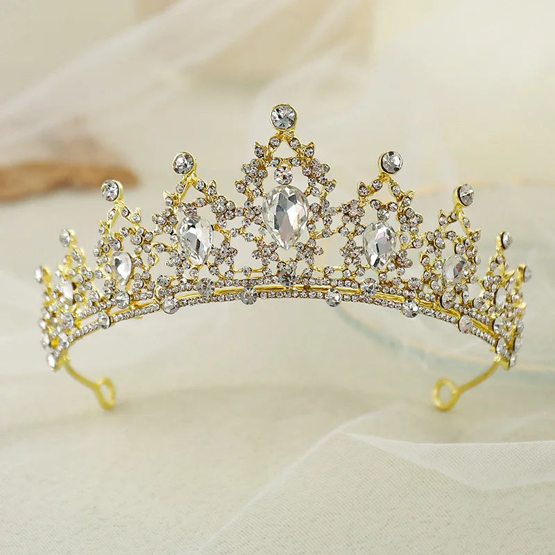 A gold tiara adorned with intricate crystal patterns and large teardrop gems, elegantly set on a soft, blurred fabric background.