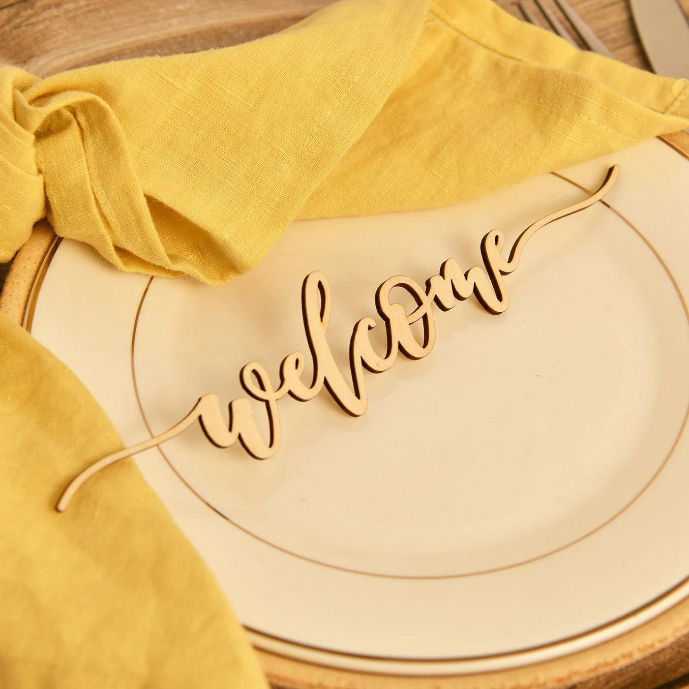 A wooden "welcome" sign rests on a white plate with a gold rim, accompanied by a folded yellow napkin, creating an inviting table setting ambiance.