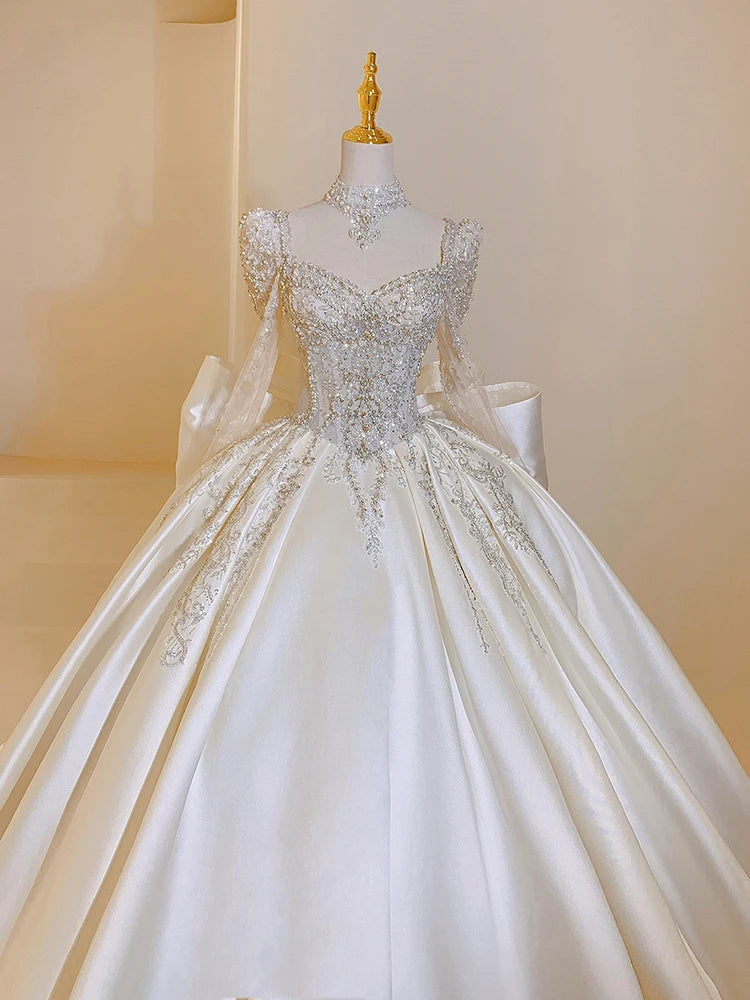Elegant white wedding dress on a mannequin with intricate silver embroidery, beaded bodice, and lace details on long sleeves. The skirt is voluminous and flowing.