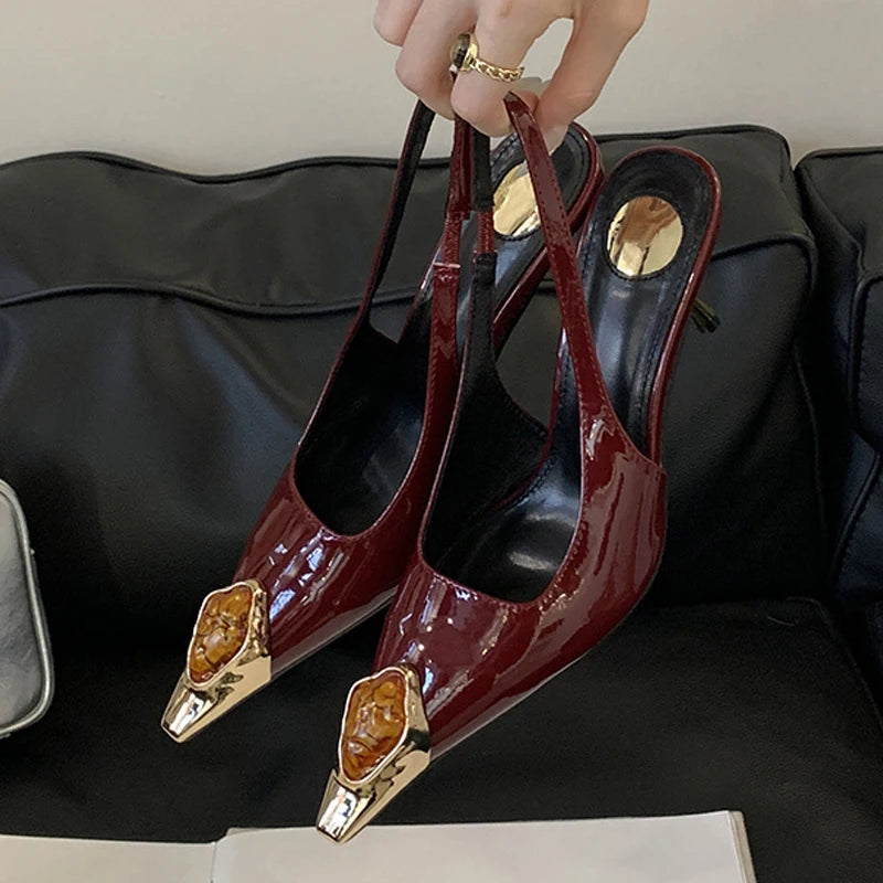 Hand holding glossy maroon slingback heels with ornate gold and amber embellishments on the pointed toes, set against a black leather background.
