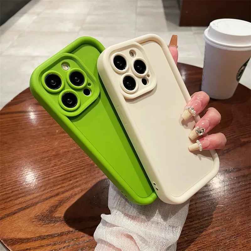 A hand with manicured nails holds two smartphones in green and beige cases with triple cameras, over a wooden table. A white disposable cup is visible in the background.