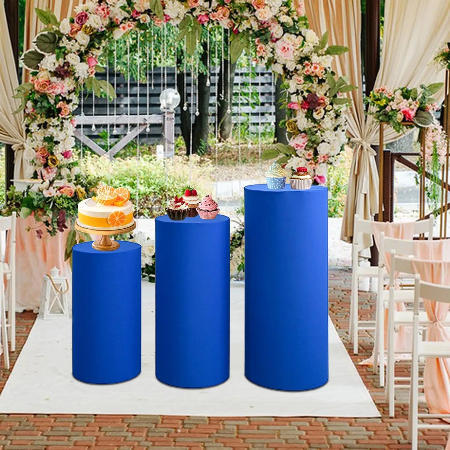 A floral wedding arch frames three blue columns displaying a citrus cake and colorful cupcakes. White chairs and draped fabric complete the outdoor ceremony setting.