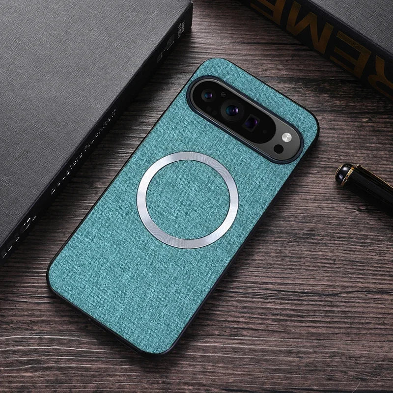 Teal fabric phone case with a metal ring for MagSafe, resting on a wood table. Nearby are a gray notebook and a black pen with gold accents.