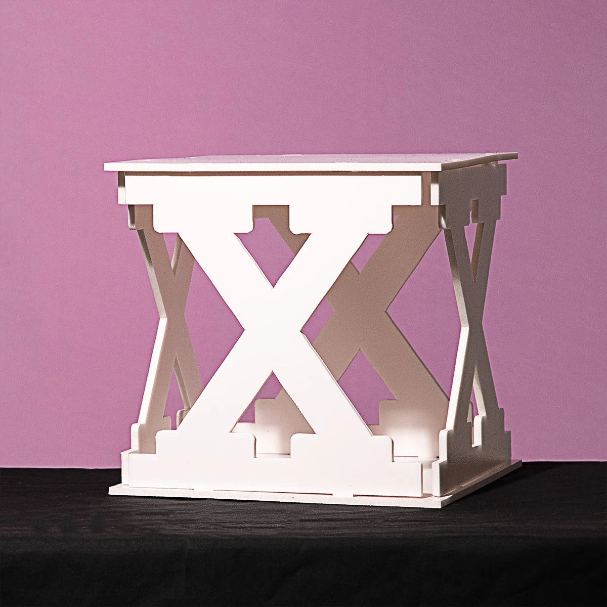 White cubic structure with intersecting cross patterns on each side, placed on a black surface against a pink background.
