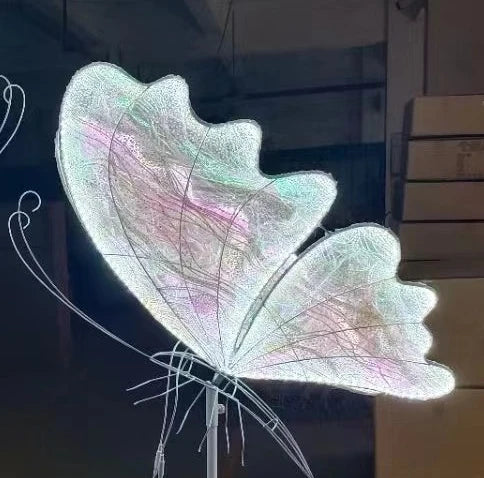 A glowing butterfly sculpture with iridescent wings displaying pink, green, and blue hues, standing on a delicate metal structure against a dark background.