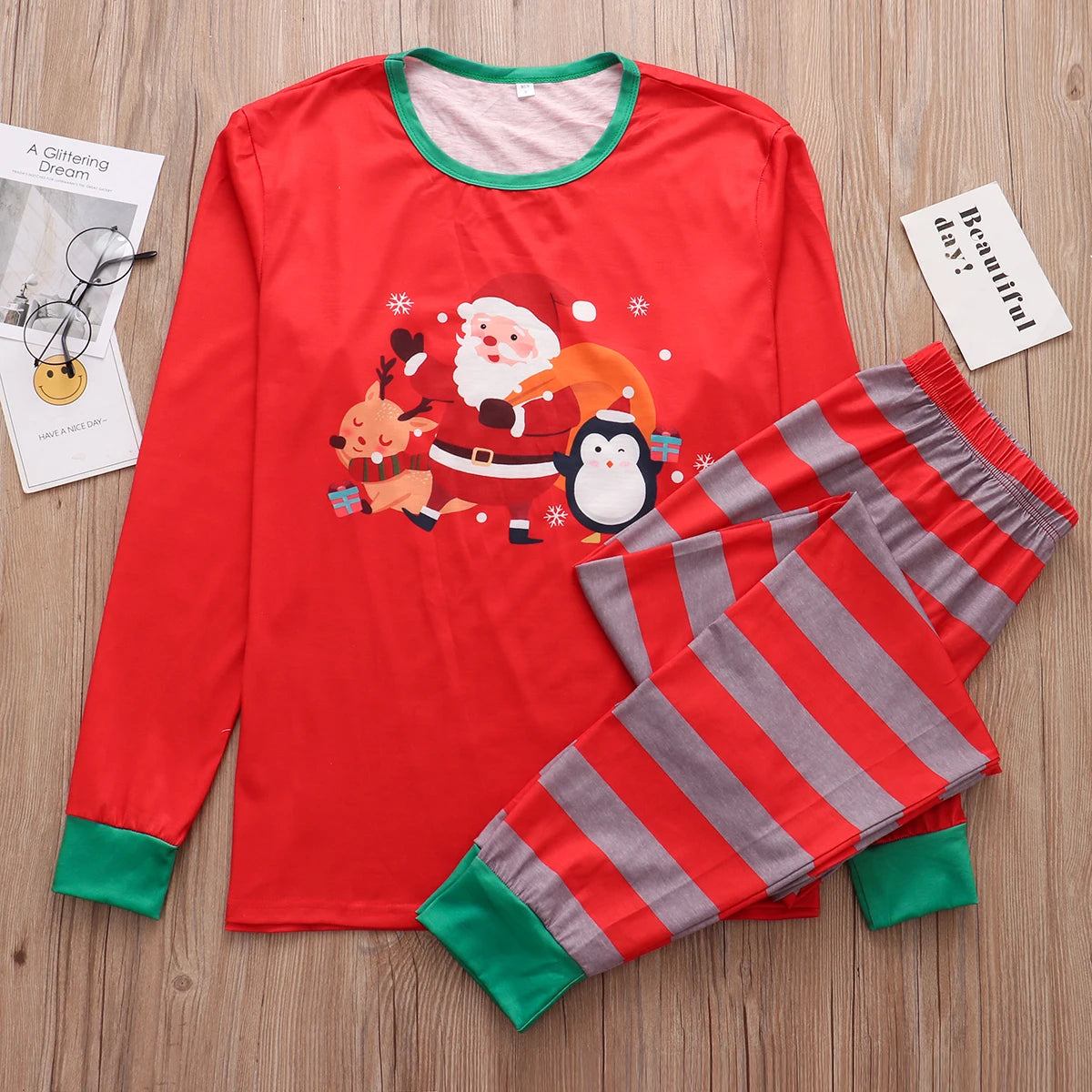 Red Christmas-themed pajamas featuring Santa, a reindeer, and a penguin. The set includes a long-sleeved top and striped pants. Glasses and booklets are placed beside on a wooden surface.