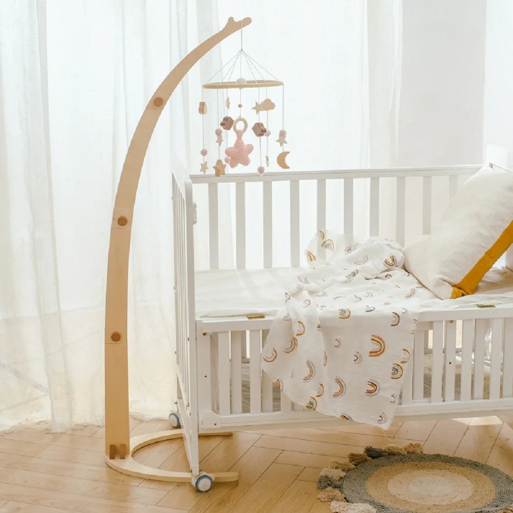 A white crib with a crescent-themed blanket, wooden mobile stand, and soft hanging toys. Light streams through sheer curtains onto a woven round rug on the wooden floor.