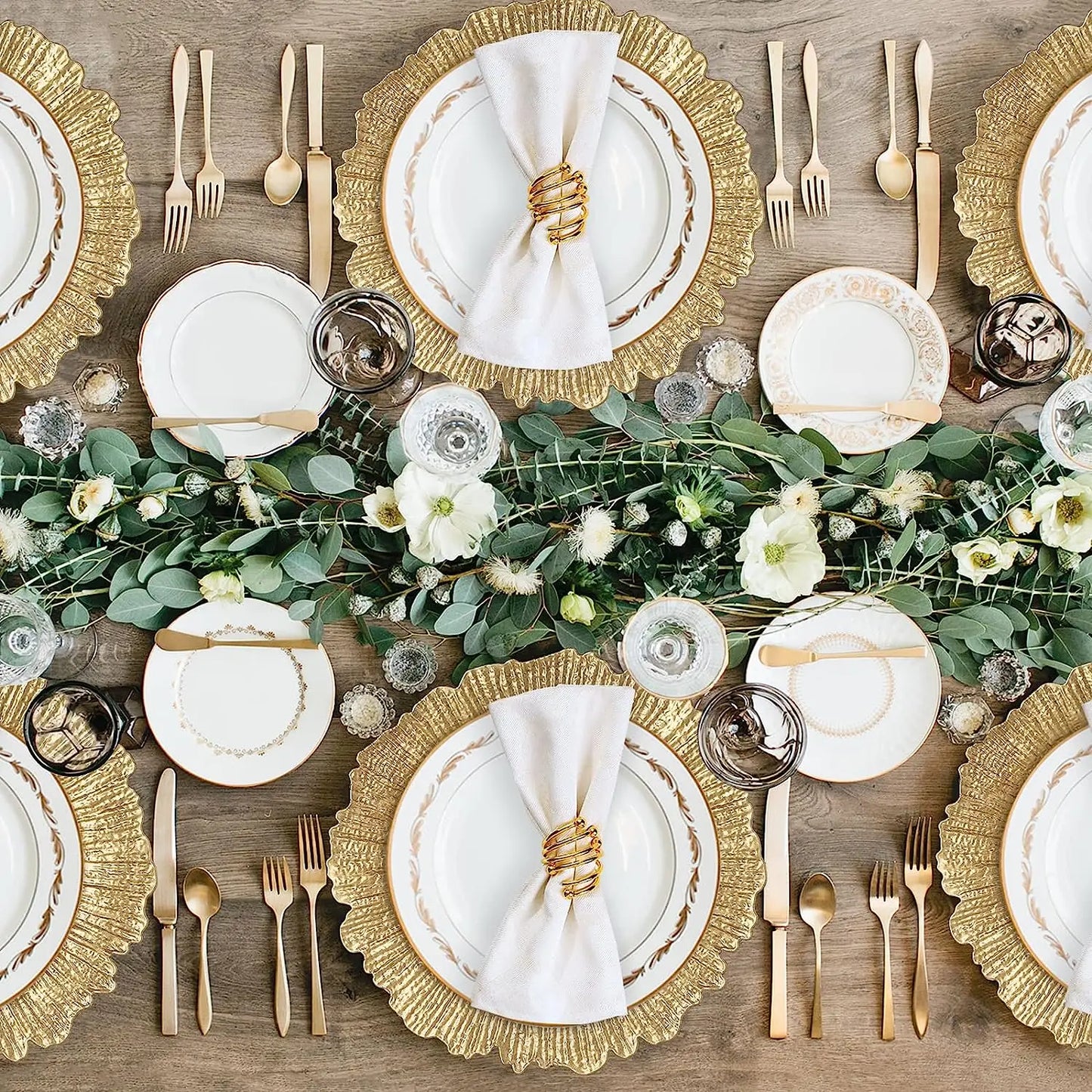 Elegant table setting with gold-rimmed plates and cutlery, white napkins in gold rings, and a lush green floral centerpiece featuring white flowers on a wooden table.