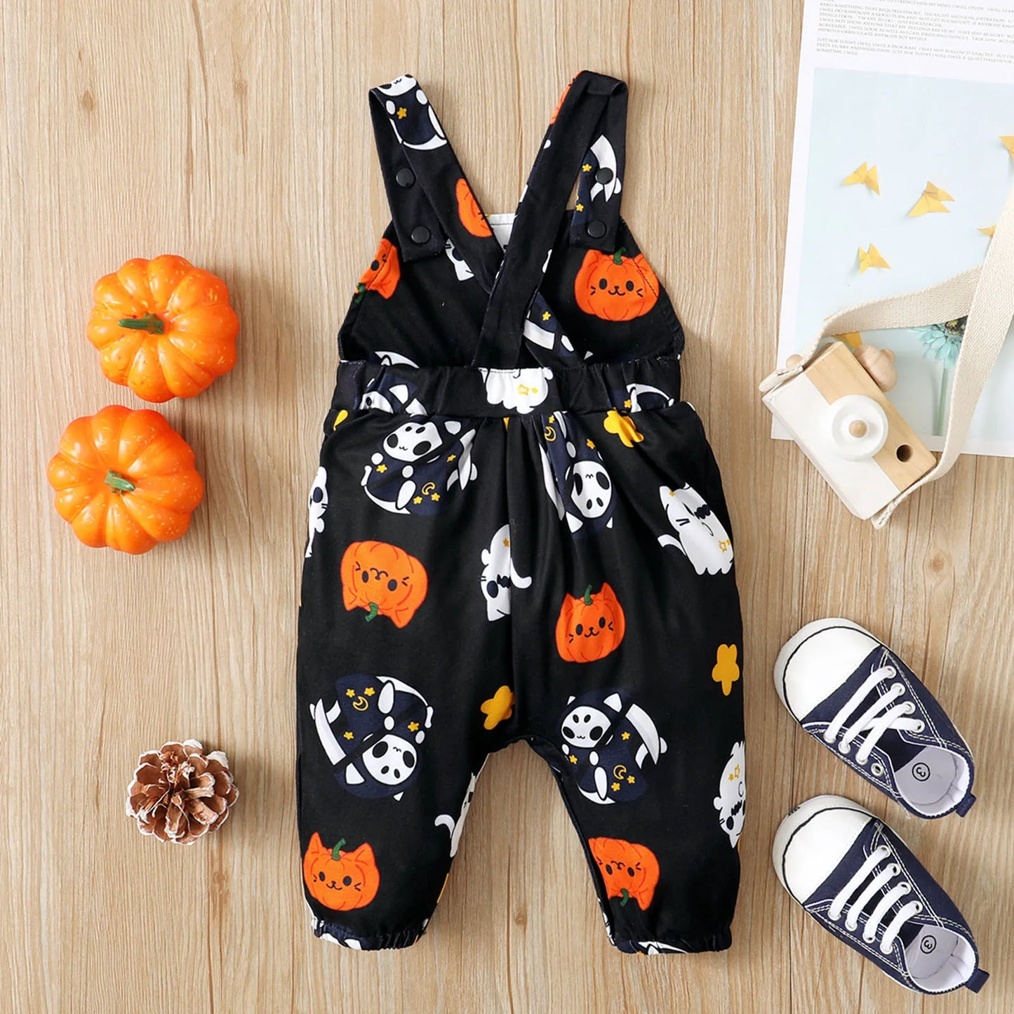 Black toddler romper with cute Halloween prints of ghosts, pumpkins, and bats. Nearby are mini pumpkins, a wooden camera, pinecone, and tiny sneakers.