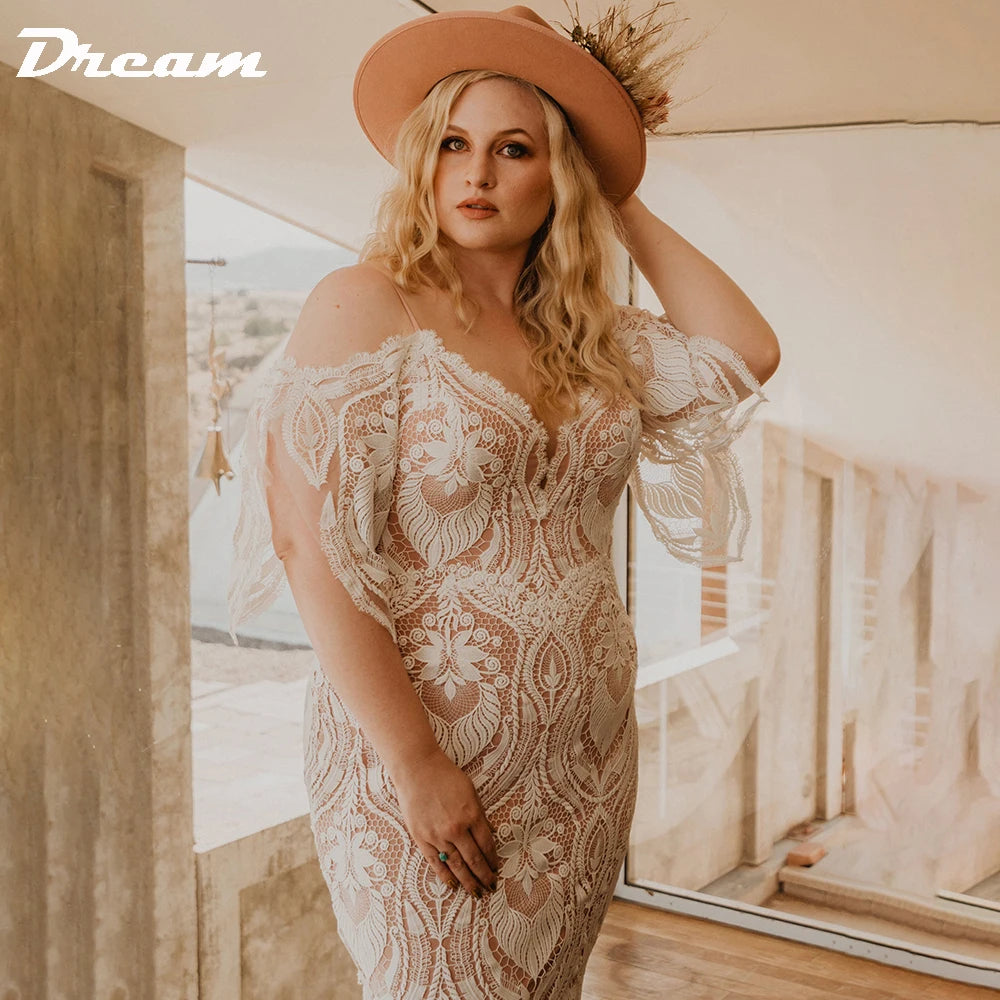 Woman in a detailed lace gown with off-shoulder sleeves poses indoors. She wears a tan hat adorned with feathers, and stands in front of a large window, creating a soft, warm ambiance.