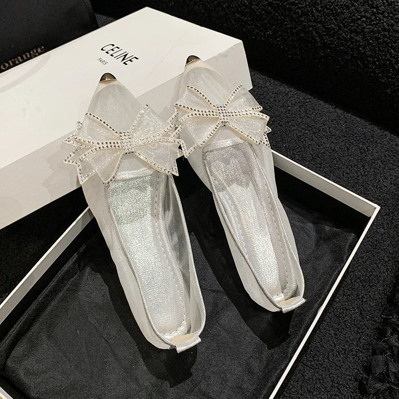 Elegant satin flats with metal-tipped toes and embellished bows, displayed in an open box. A branded shoe box is partially visible in the background.