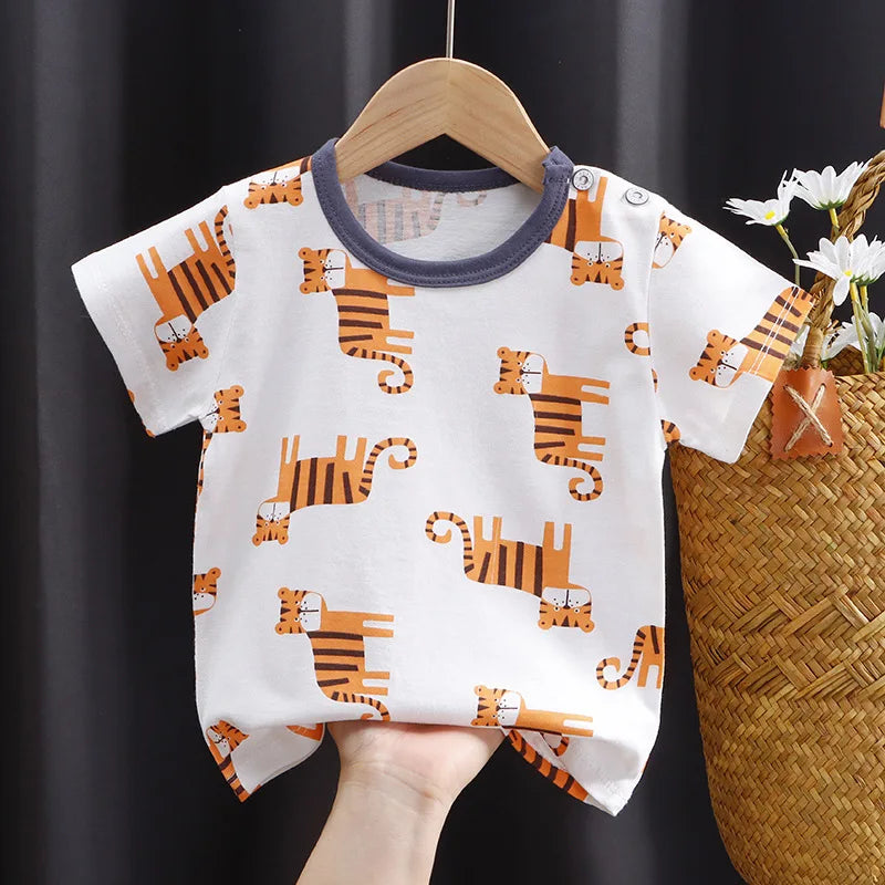 White baby onesie with orange tiger print, hanging on a wooden hanger. A hand holds the bottom. A woven basket with white daisies is partially visible on the right.