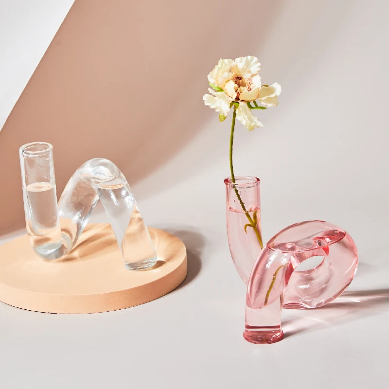 Two modern, curved glass vases, one clear and one pink. The pink vase holds a single white flower, creating an artistic display against a soft, neutral background.