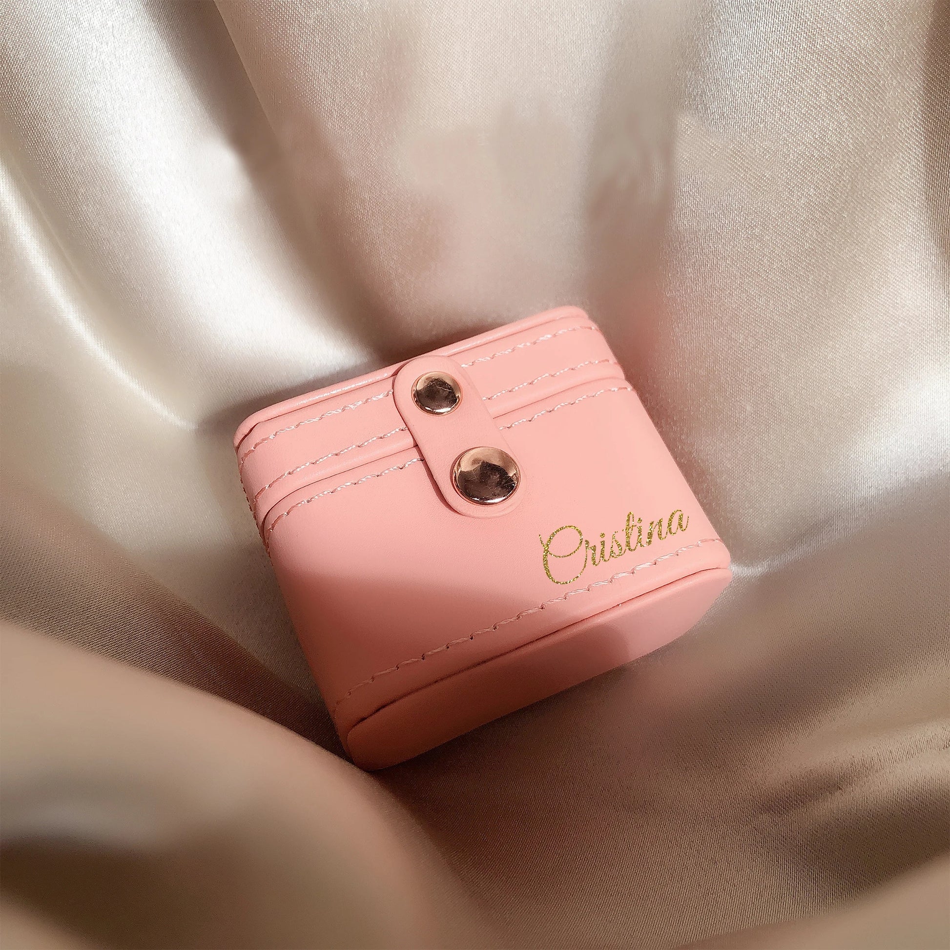 Delicate pink leather box with two metal studs, personalized with the name "Cristina" in gold script, rests on luxurious satin fabric with soft, flowing folds.