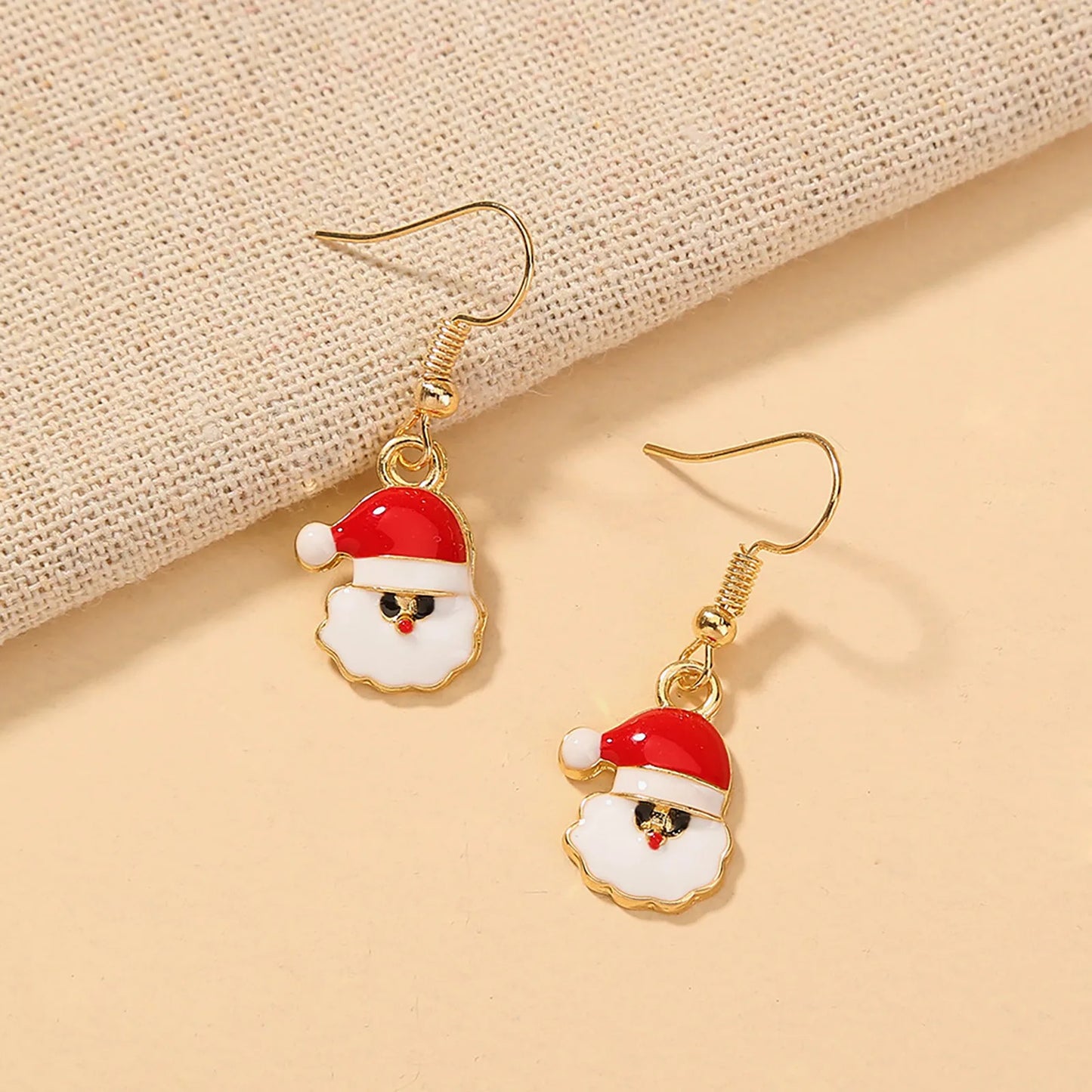 Golden hook earrings featuring Santa Claus faces with red hats and white beards, set on a light, textured fabric background.