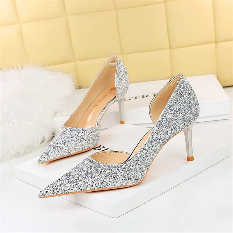 Sparkling silver stiletto heels with pointed toes, resting on white boxes. The shoes have a glittery texture and are set against a soft, quilted yellow background.
