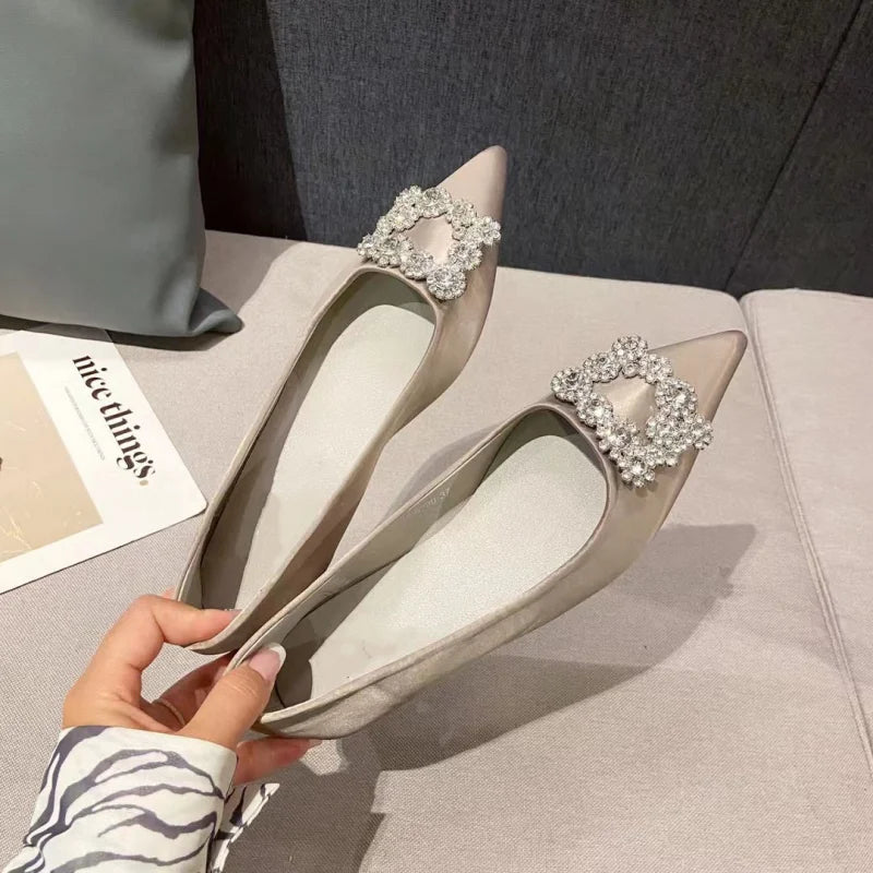 Elegant, pointed-toe beige flats with sparkling floral embellishments on the toes, held by a hand over a light pink sofa near a magazine titled "nice things."