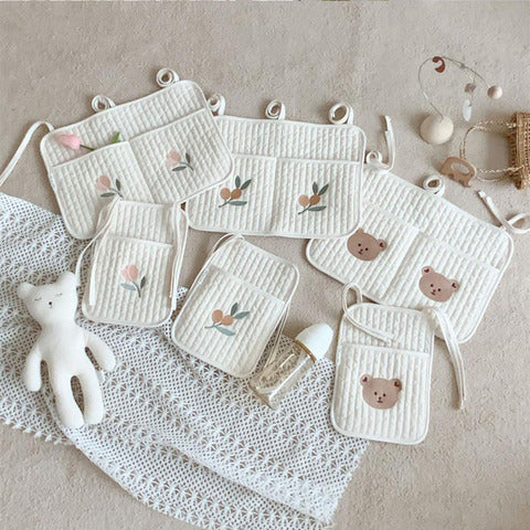 Assorted baby bibs with floral and bear designs, neatly arranged on a lace cloth. Nearby, a white plush bear, a baby bottle, and a hanging decoration with a wooden ring.