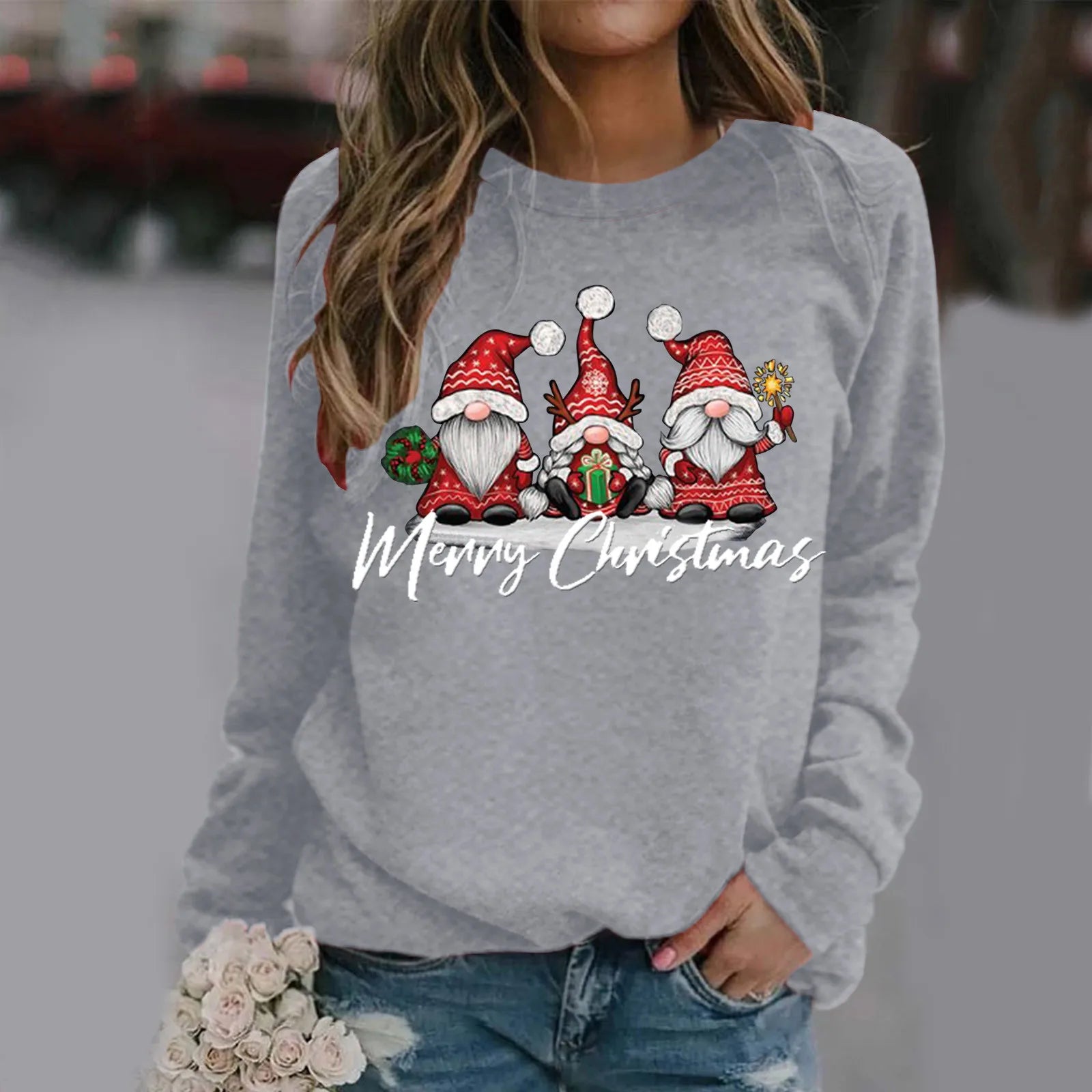 Woman in a grey sweater with cartoon Christmas gnomes and "Merry Christmas" text, holding a small bouquet of roses. Jeans complement the casual look.