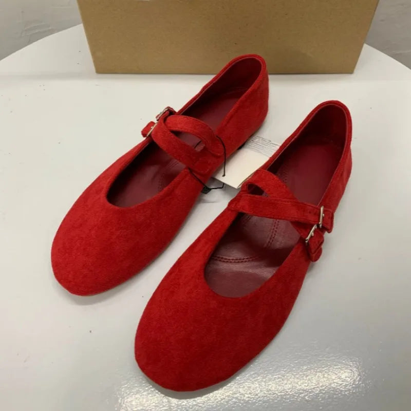 Pair of red suede flats with buckle straps, displayed on a white surface. The shoes are elegant, with rounded toes, and sit in front of a plain brown box.