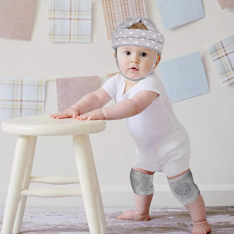 A baby wearing a padded helmet and knee pads stands using a small stool for support. The background features pastel-patterned fabric squares on the wall.