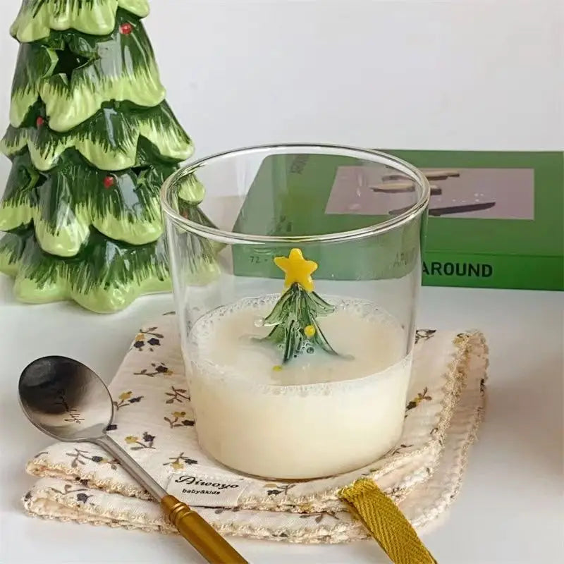 A glass of milk with a Christmas tree decoration inside, placed on a floral napkin. A green ceramic tree and a green box are in the background, with a spoon nearby.
