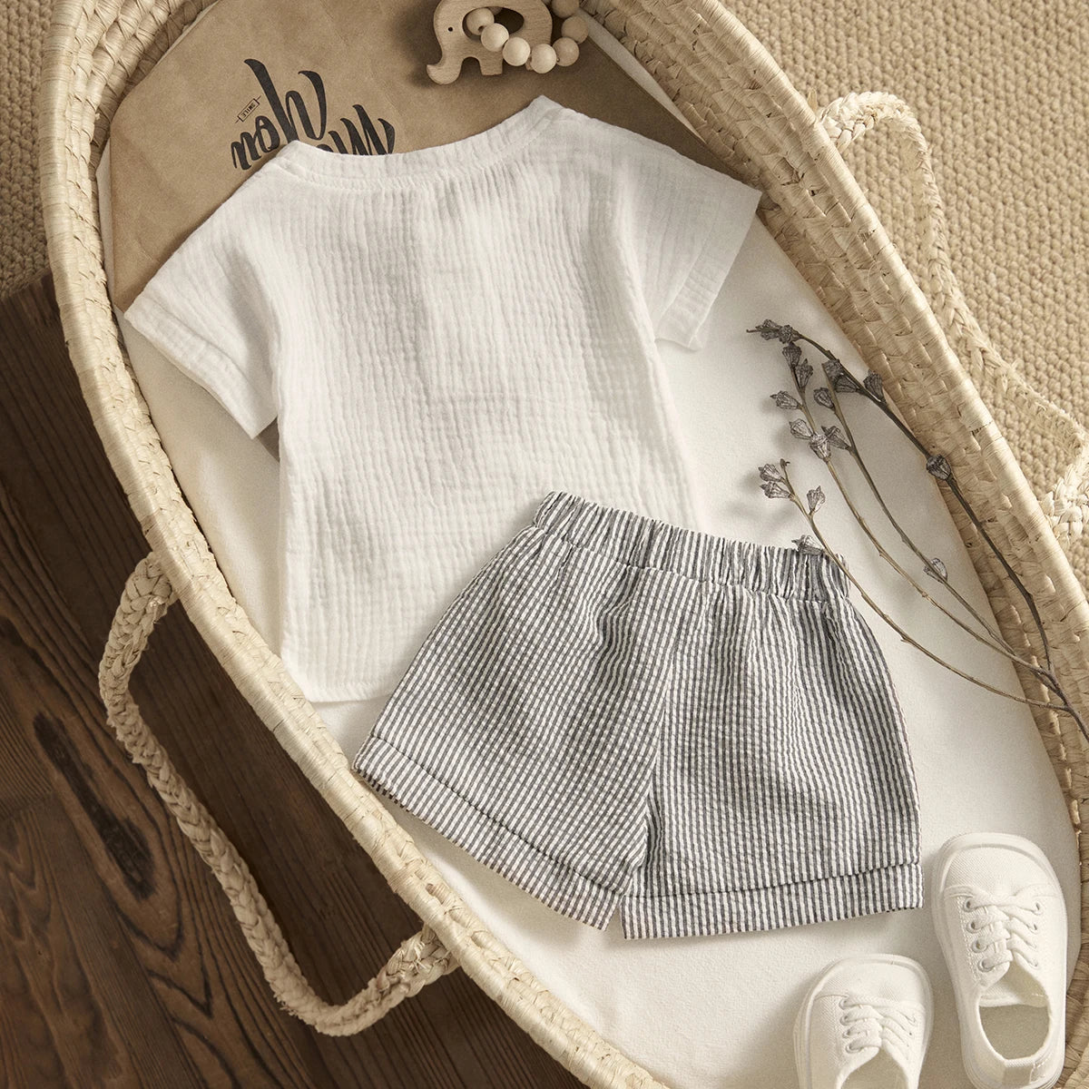 Baby outfit laid in a woven basket: white textured short-sleeve shirt, gray striped shorts, white baby shoes. Dried flowers and wooden elephant toy add charm.