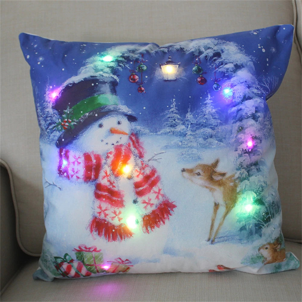 Festive pillow with a snowman in a top hat and scarf, a deer, and gifts in a snowy scene, illuminated by colorful LED lights.