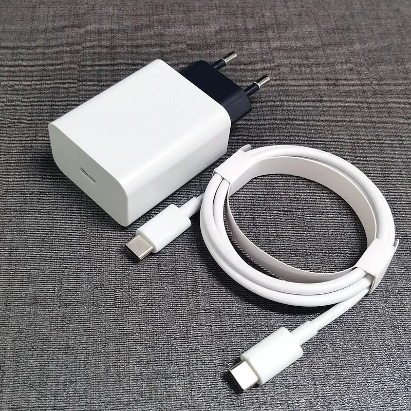 White USB-C charging block and cable on a gray textured surface. The block has two round pins and a USB-C port. The cable is coiled neatly with USB-C connectors.