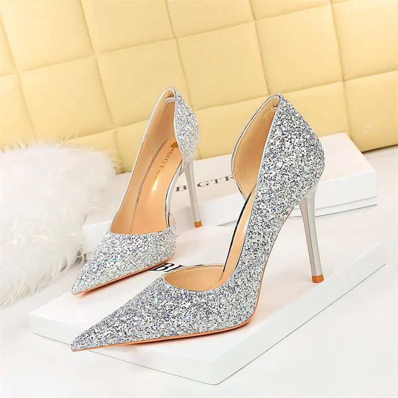 Sparkling silver stiletto heels on a white surface, featuring a pointed toe design. The shoes are set against a soft, yellow quilted background, with a fluffy decor nearby.