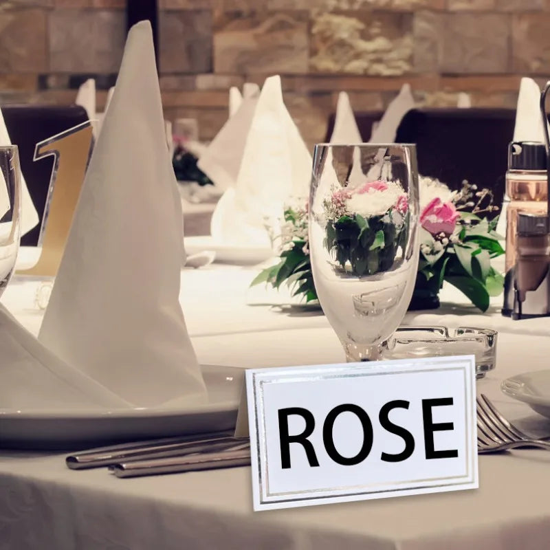 Elegant table setting with white napkins, wine glass, floral centerpiece, and a name card labeled "ROSE" in focus, against a warm-toned, blurred background.