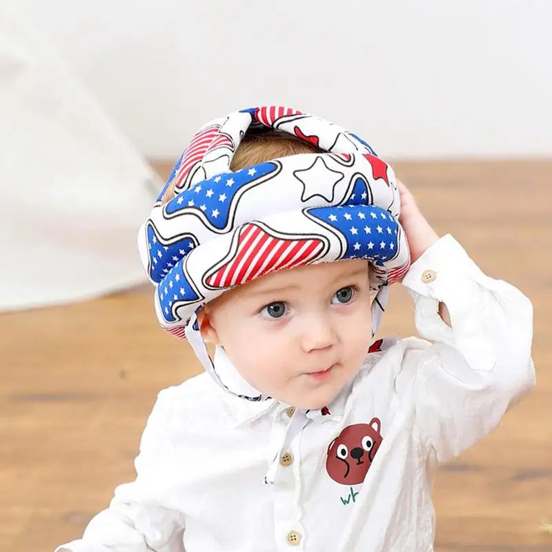 A young child wearing a white shirt with a bear design and a padded helmet featuring red, white, and blue stars and stripes sits on a wooden floor, looking curious.