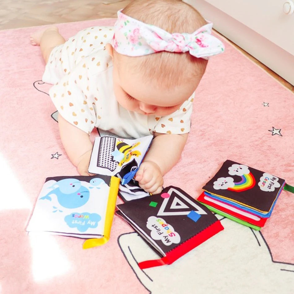 A baby lying on a pink mat, wearing a floral headband and heart-patterned outfit, plays with cloth books featuring colorful designs and shapes.