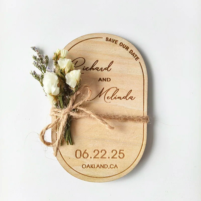 Wooden save-the-date card with "Richard and Melinda," adorned with white flowers tied with twine. Date 06.22.25 and location Oakland, CA, are engraved.