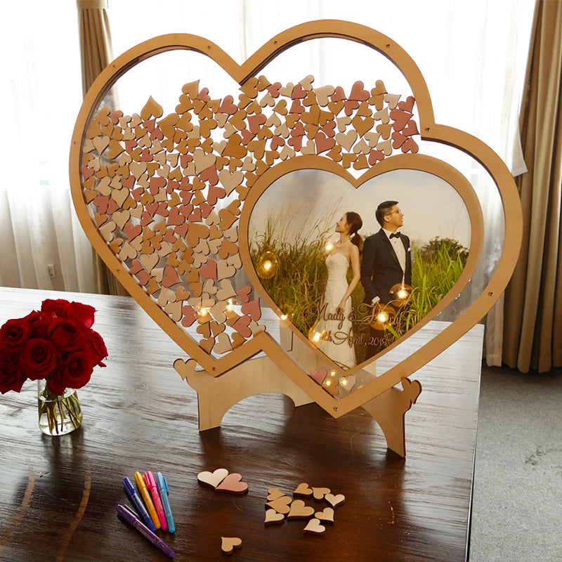 Wedding guestbook with interlocking hearts and couple's photo inside sits on a table. Red roses, pens, and heart-shaped pieces are beside it.