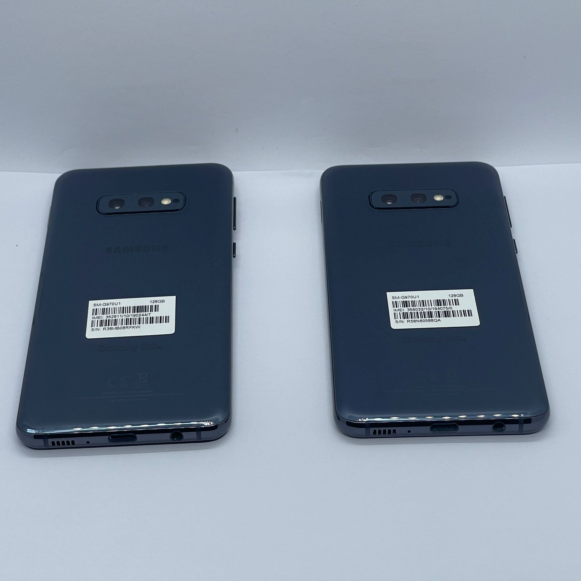 Two Samsung Galaxy S10e smartphones, back view, placed side by side. Each has dual rear cameras and visible product labels with barcodes.