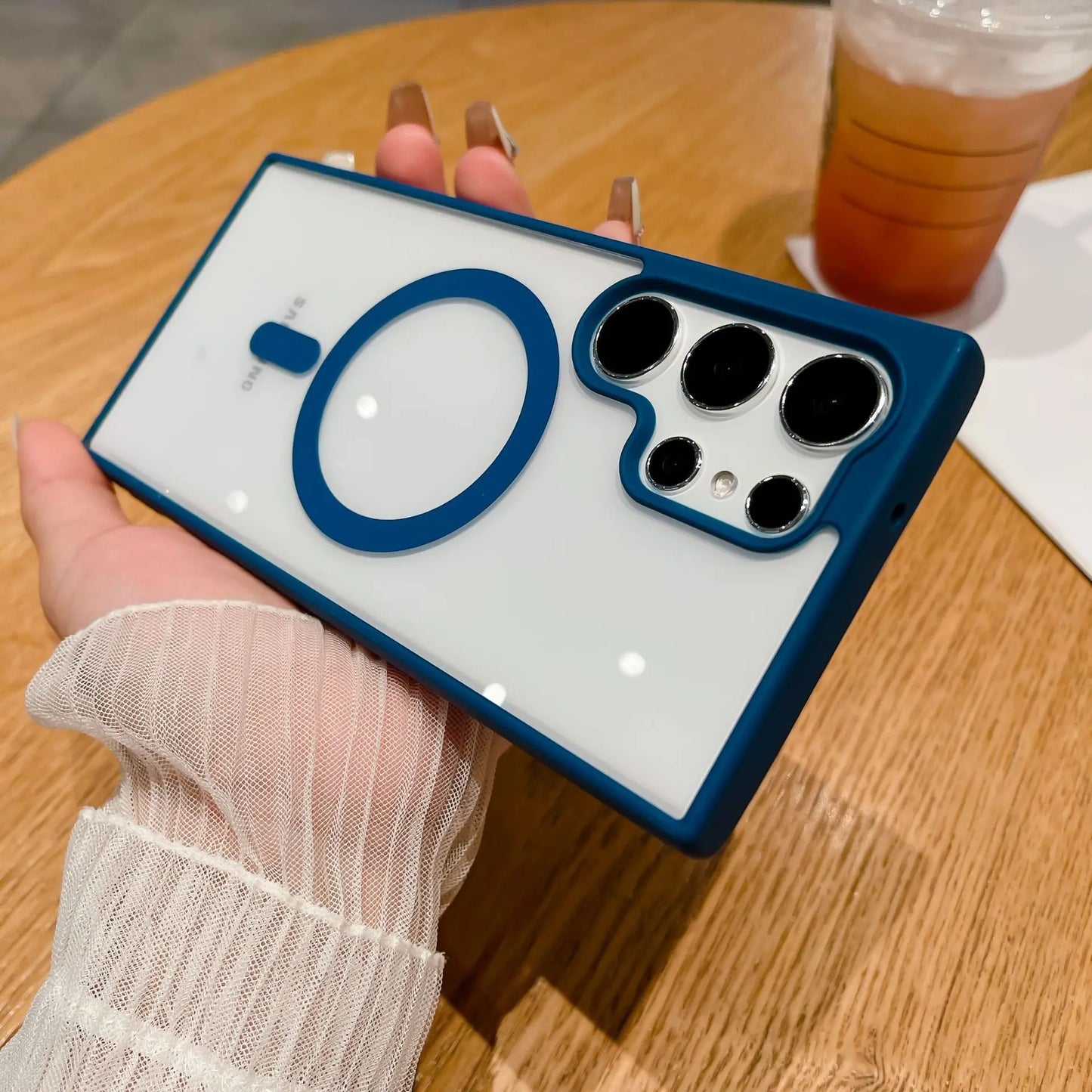 A person holds a phone with a blue and white case featuring a large circle design. The camera has multiple lenses. A drink with ice is on a wooden table in the background.