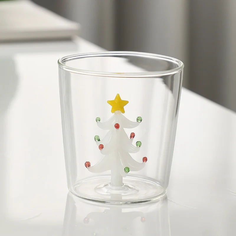 Clear glass cup featuring a white Christmas tree with red and green ornaments inside, topped by a yellow star, placed on a reflective white surface.