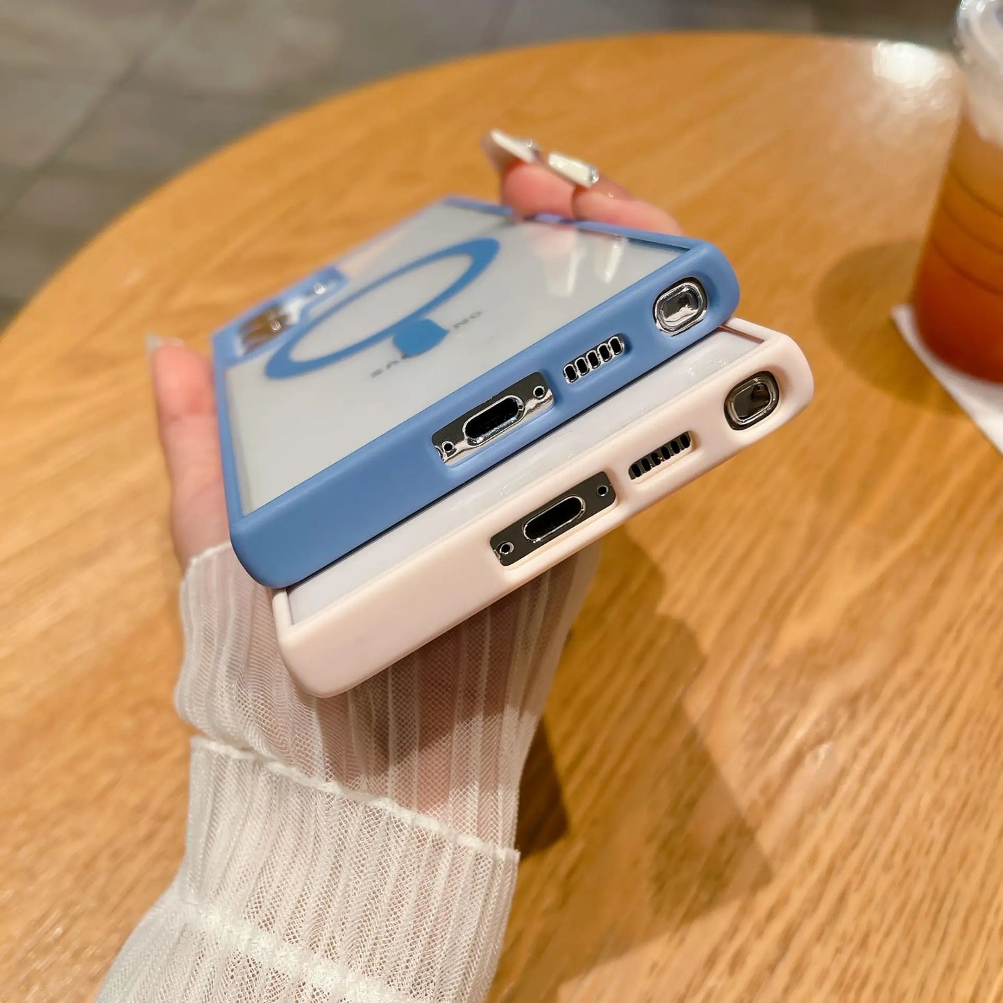 Two smartphones with camera lenses and charging ports visible are held in hand. One has a blue case, the other white. A drink sits on a wooden table nearby.