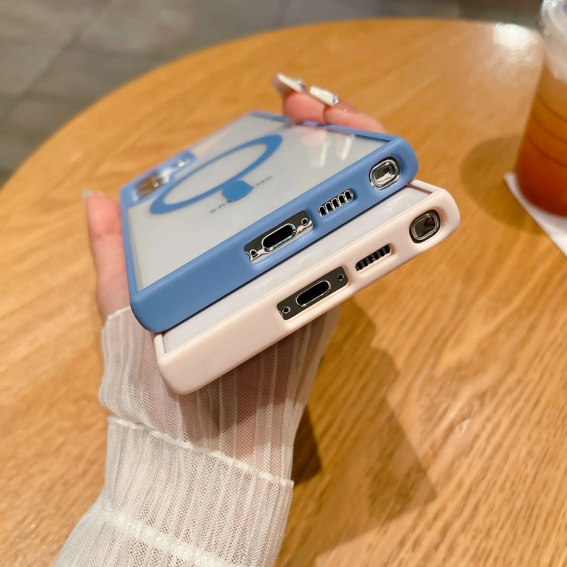 Two smartphones with camera lenses and charging ports visible are held in hand. One has a blue case, the other white. A drink sits on a wooden table nearby.