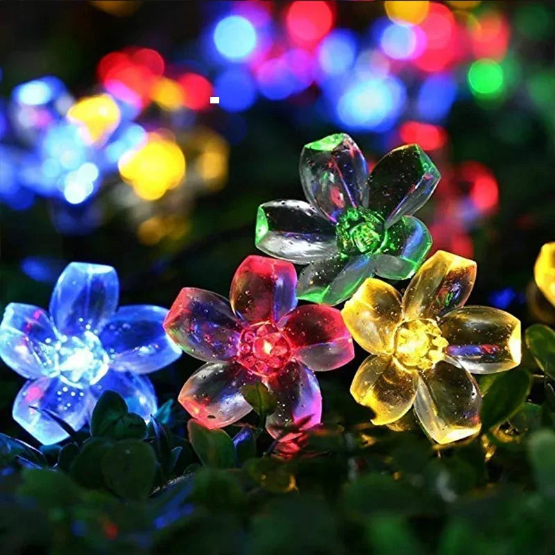 Colorful LED flower lights in purple, red, yellow, and green are surrounded by dark leaves. The lights create a glowing, festive atmosphere against a blurred background.
