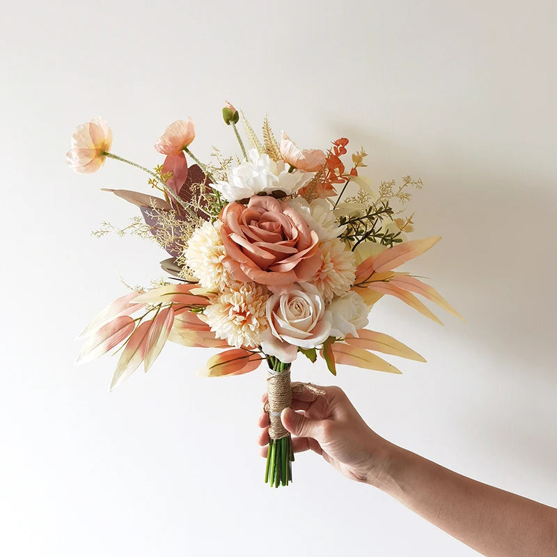 Hand holding a bouquet with soft pink, white, and peach flowers, including roses and peonies, accented by delicate greenery. The background is a plain white wall.