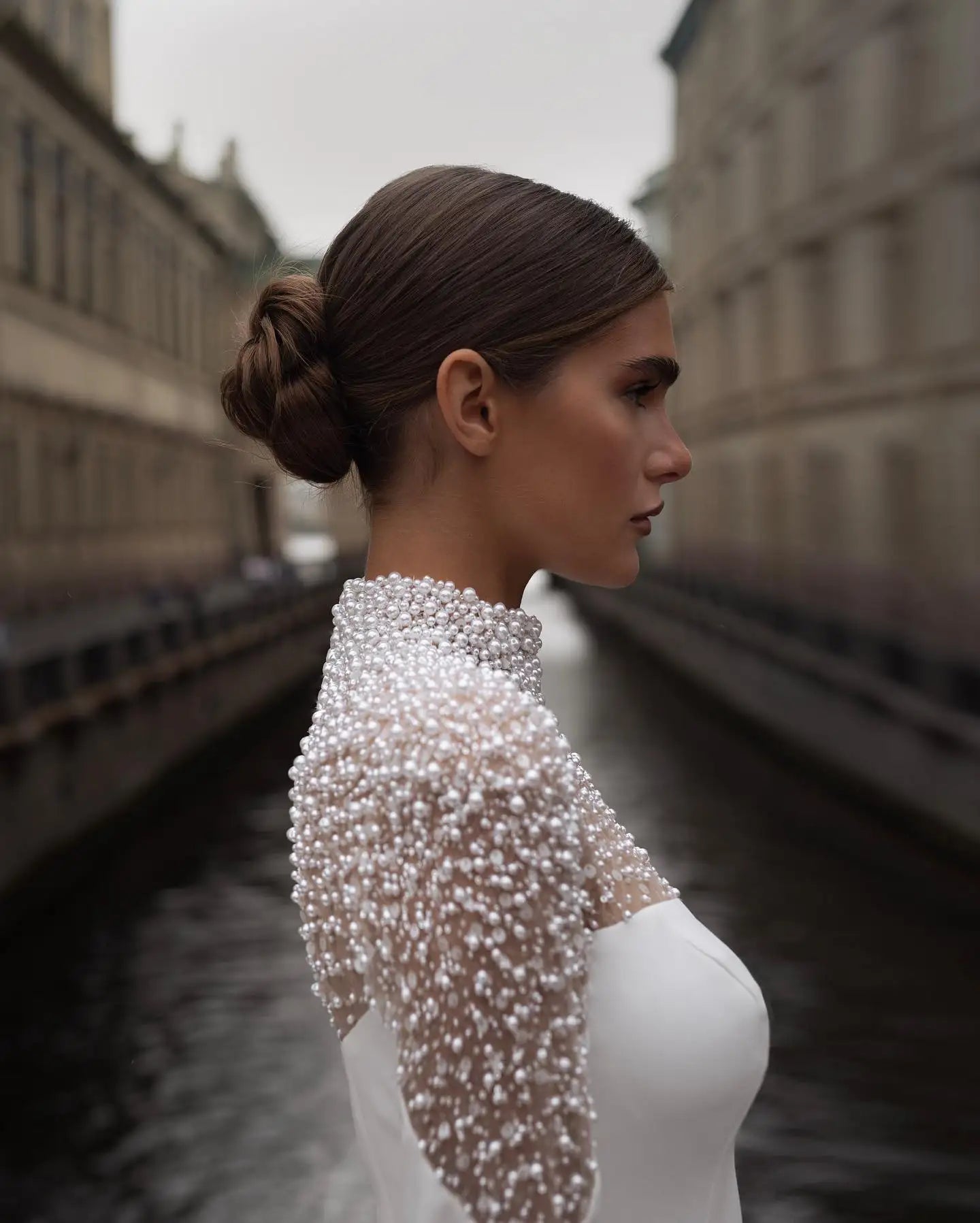 A woman in a pearl-embellished, high-neck dress stands in profile against a blurred cityscape with water and historic buildings, her hair elegantly tied back.