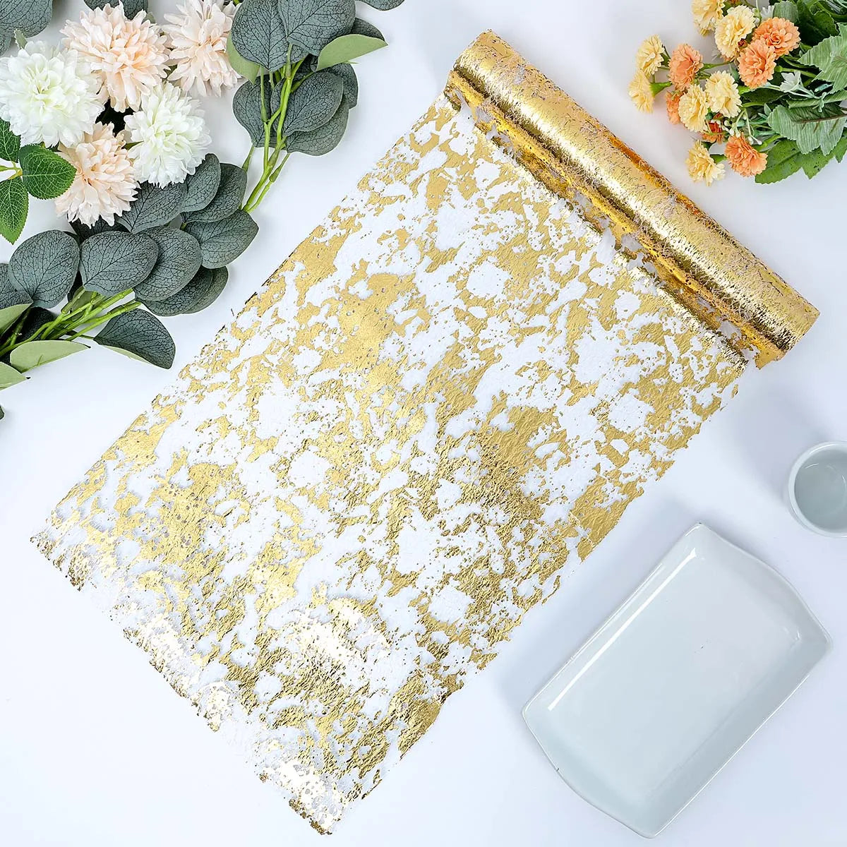 Gold and white textured wallpaper roll on a table, surrounded by green foliage and pastel flowers. Nearby are a white rectangular plate and a small cup on the white surface.